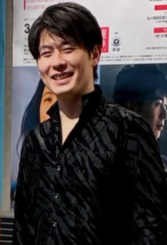 黒いシャツを着用し、笑顔でカメラを見る福間洸太朗。背景にポスター。