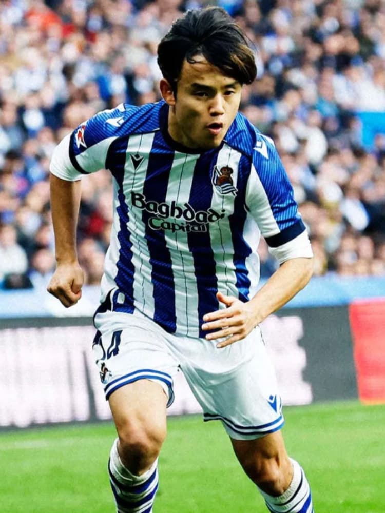 ピッチ上でレアル・ソシエダの青と白のストライプユニフォーム（Baghdad Capitalロゴ）を着用して走る、中距離ショットの久保建英。