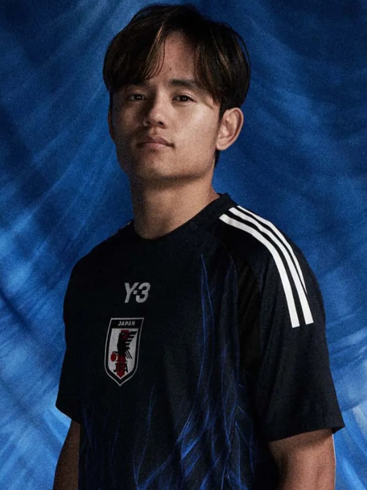 JFAのエンブレムとY-3ロゴが入った黒い日本代表練習着を着用し、正面を見つめる久保建英のクローズアップ。青い抽象的な背景。