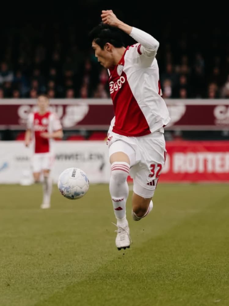 アヤックスの2025-26シーズンホームキット（背番号32）を着用し、ピッチ上で空中にあるボールを胸でコントロールしようとする冨安健洋のアクション写真。