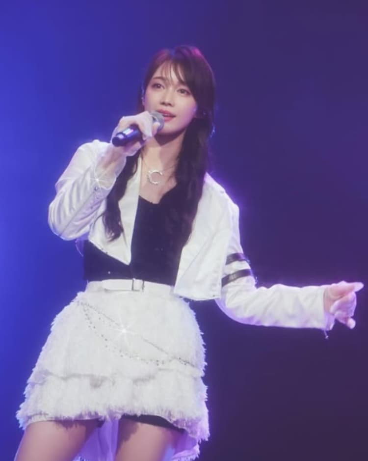 ステージ上でマイクを握り、スポットライトを浴びて歌唱するアーティスト・中村ゆりか。白いフェザー調のミニスカートにクールなジャケットを合わせた衣装をまとい、表現者としての悦びに満ちた表情を見せている。