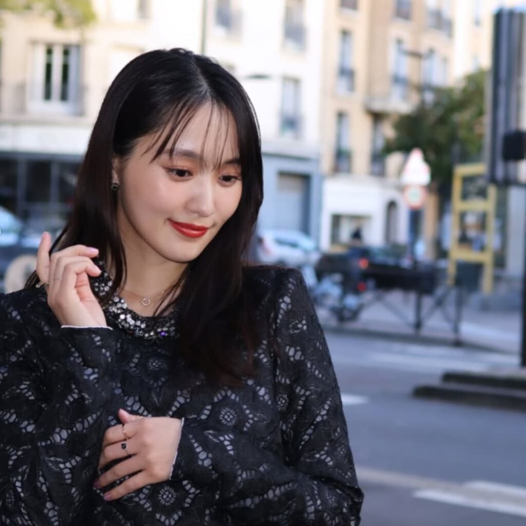 黒いレースのワンピースを着用し、赤いリップが印象的なメイクでヨーロッパ風の街並みを背景に視線を落として佇む菅井友香。