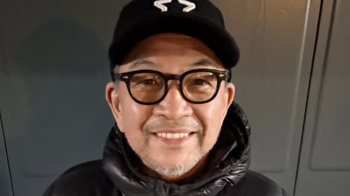 眼鏡と黒いダウンジャケット、白いロゴの入った黒い野球帽を着用した中野英雄氏が、グレーのシャッターを背景に、カメラに向かって微笑んでいるアップ写真。