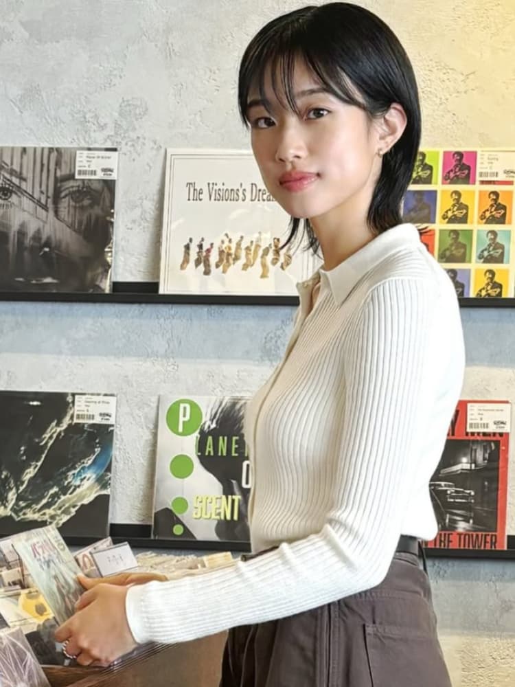 レコードショップで、白いリブニットカーディガンを着用しショートヘアでレコードを手に取る河合優実。背景には多数のレコードが並んでいる。