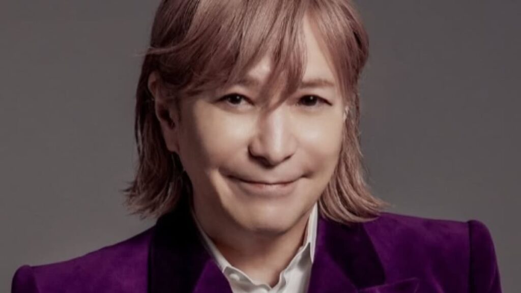 2026年現在の小室哲哉氏のポートレート。落ち着いた赤茶色のミディアムヘアに、鮮やかな紫色のジャケットと白いシャツを纏い、穏やかな微笑みを浮かべているアーティスト写真。