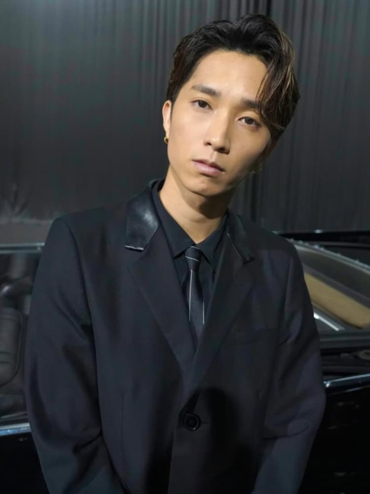 黒髪の上半身ショット。田中樹は黒いスーツに黒いネクタイを着用し、ゴールドのフープイヤリングをつけている。カメラを真剣に見つめている。背景は暗く、黒い車の一部が見える。