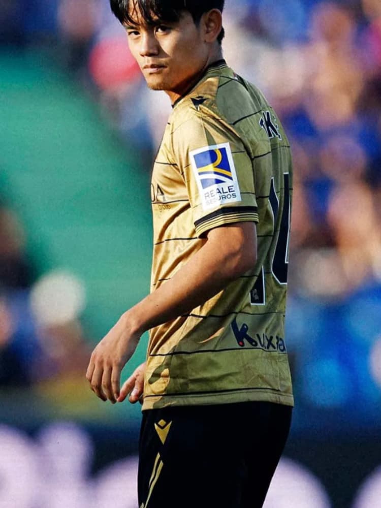 レアル・ソシエダのゴールドのユニフォーム（Reale Segurosロゴ）を着用し、肩越しに振り返る後ろ姿の久保建英のクローズアップ。