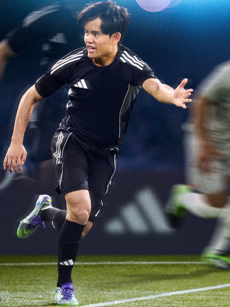 ピッチ上で黒い練習着を着用し、ボールに向かって突進する久保建英の全身ショット。低いアングル。