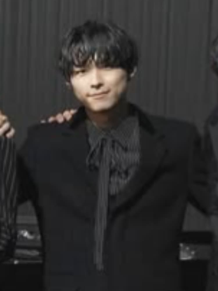 松村北斗氏。image_1.pngと同じ。黒い波打った髪。カメラを真っ直ぐ見つめている。落ち着いた表情。image_1.pngと同じ。黒いスーツジャケットの下に、黒と白のストライプシャツを着用。image_1.pngよりも少し引きのショット。背景は暗い、フォーマルな場所。