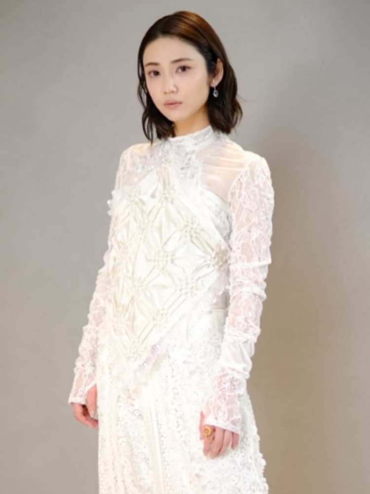 暗いボブヘアで自然な表情の山谷花純が、全身白の繊細なレースドレスを着て立っているポートレート。ドレスはハイネックで、胸元に装飾がある。背景はニュートラルなグレー。