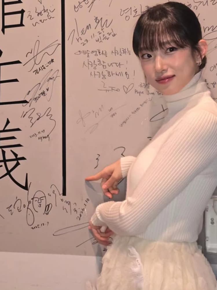 サインが多数書かれた壁の前に立ち、笑顔で壁を指差す白いタートルネックニットとチュールスカート姿の河合優実。