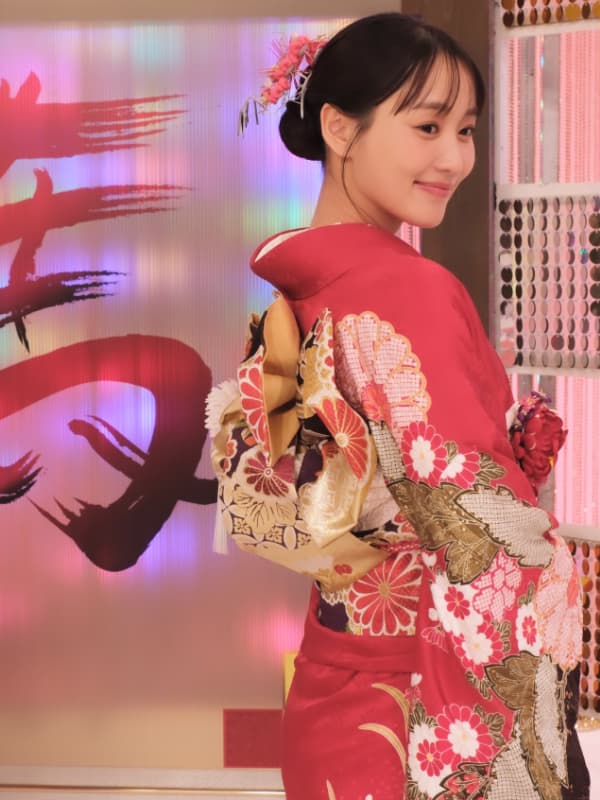 華やかな赤い振袖を着用し、花の髪飾りをあしらったまとめ髪で優しく振り返る菅井友香の姿。背景には「寿」の文字が書かれた和風の装飾がある。