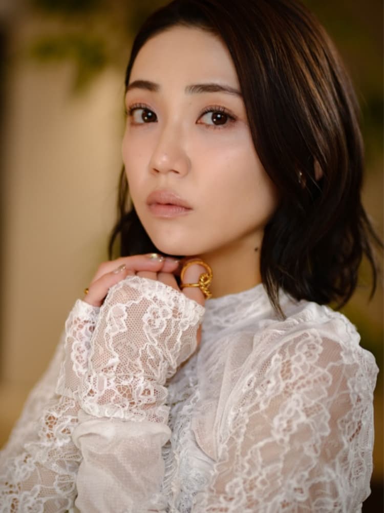 暗いボブヘアの山谷花純が、顎の下で手を重ねてカメラを見つめるクローズアップ写真。白のレースドレスと複数のゴールドリングを着用している。背景は暖かくぼやけている。