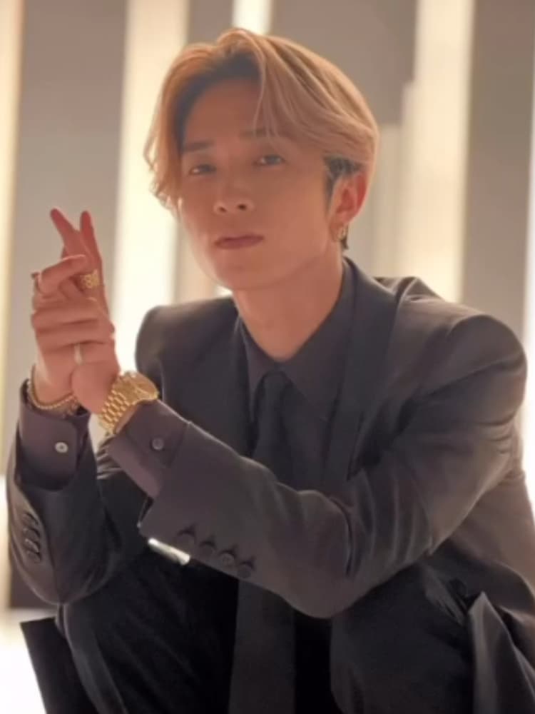 金髪の田中樹のショット。黒いスーツに黒いネクタイを着用し、ゴールドの時計と指輪を着用している。しゃがんで、両手を合わせて指を少し立てている。カメラを見つめている。背景は明るいグレーのストライプ壁。