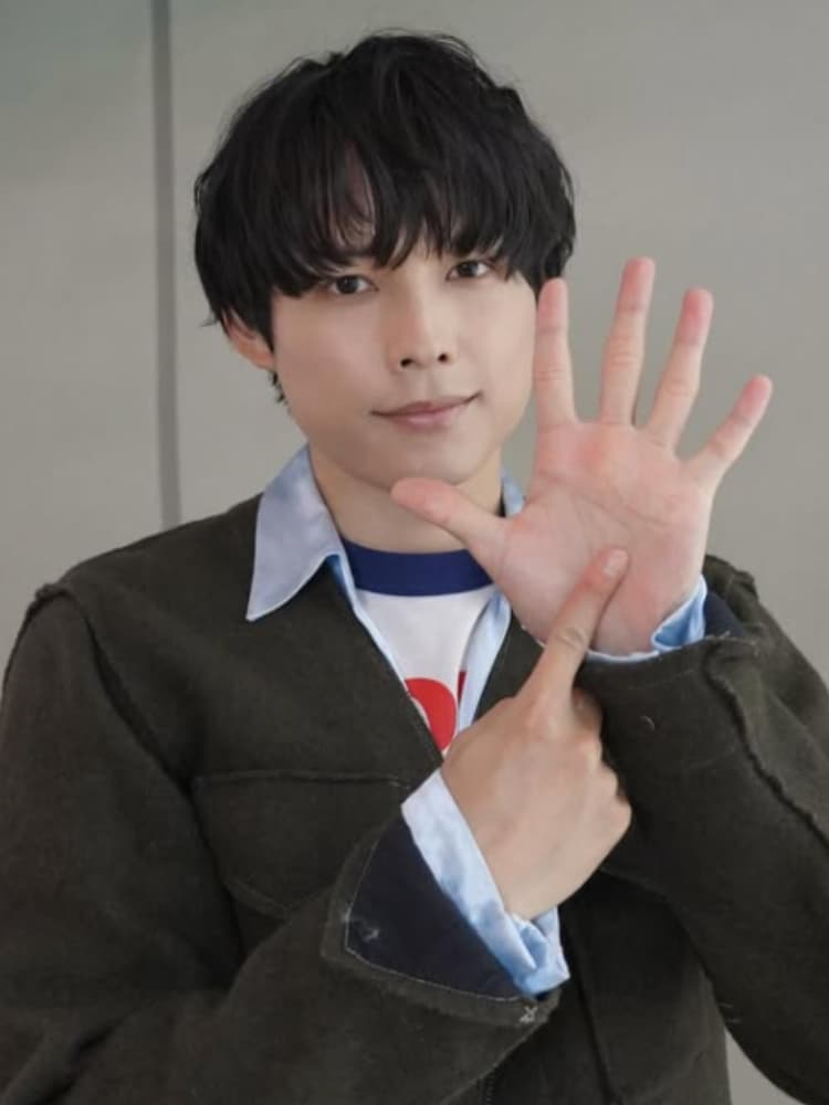 松村北斗氏。黒い髪。少し微笑んでおり、親しみやすい表情です。濃いオリーブグリーンのカーディガンの下に、ライトブルーの襟付きシャツ、さらにその下に、赤いロゴが見える白いTシャツを着用しています。両手を使い、片手（右手）で「5」の数字を作り、もう片手（左手の人差し指）でその手のひらを指して「6」の数字を作っている。これはSixTONESの「6」を表すポーズとして知られている。