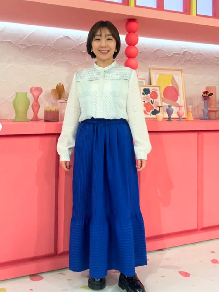白いブラウスに鮮やかな青色のロングスカートを合わせ、華やかな装飾が施されたスタジオに立つ高橋成美さんの全身ショット。