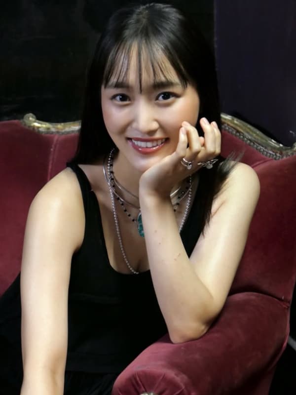 黒いノースリーブのドレスに重厚感のあるネックレスを合わせ、エンジ色のベルベットソファに座りながら顎に手を添えて微笑む菅井友香。