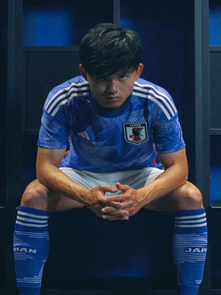 日本代表の青いユニフォームを着用し、ロッカールームで座って手を組み、真剣な表情を見せる久保建英のクローズアップ。低いアングル。