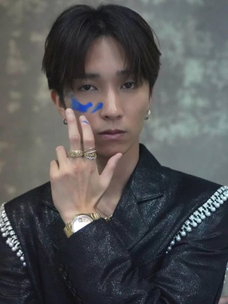 黒髪のミディアムショット。田中樹は黒いラメのジャケットを着用し、パールのような装飾が肩にある。ゴールドの時計と指輪を着用している。顔の右頬に青いペイントが施されており、右手でそれを指している。カメラを真剣に見つめている。背景は暗いグレーのテクスチャ壁。