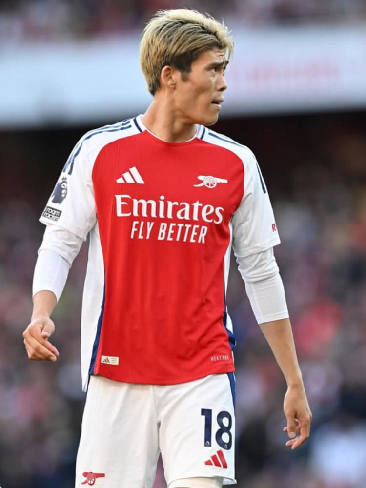 アーセナルの2024-25シーズンホームキット（赤と白、背番号18）を着用し、金髪でピッチ上に立つ冨安健洋の立ち姿。