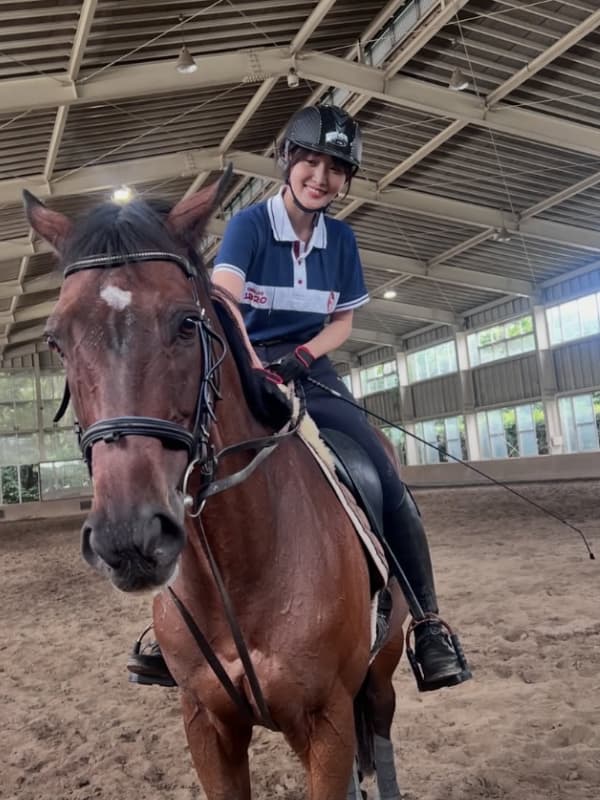 乗馬用のヘルメットと紺色のポロシャツを着用し、屋内馬場で茶色の馬に跨りながら満面の笑みを浮かべる菅井友香。