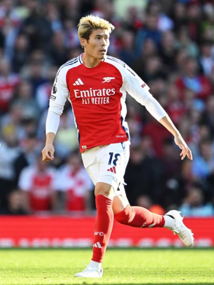 アーセナルの2024-25シーズンホームキット（背番号18）を着用し、金髪でボールをドリブルしてピッチ上を走る冨安健洋の全身写真。背景に観客席。