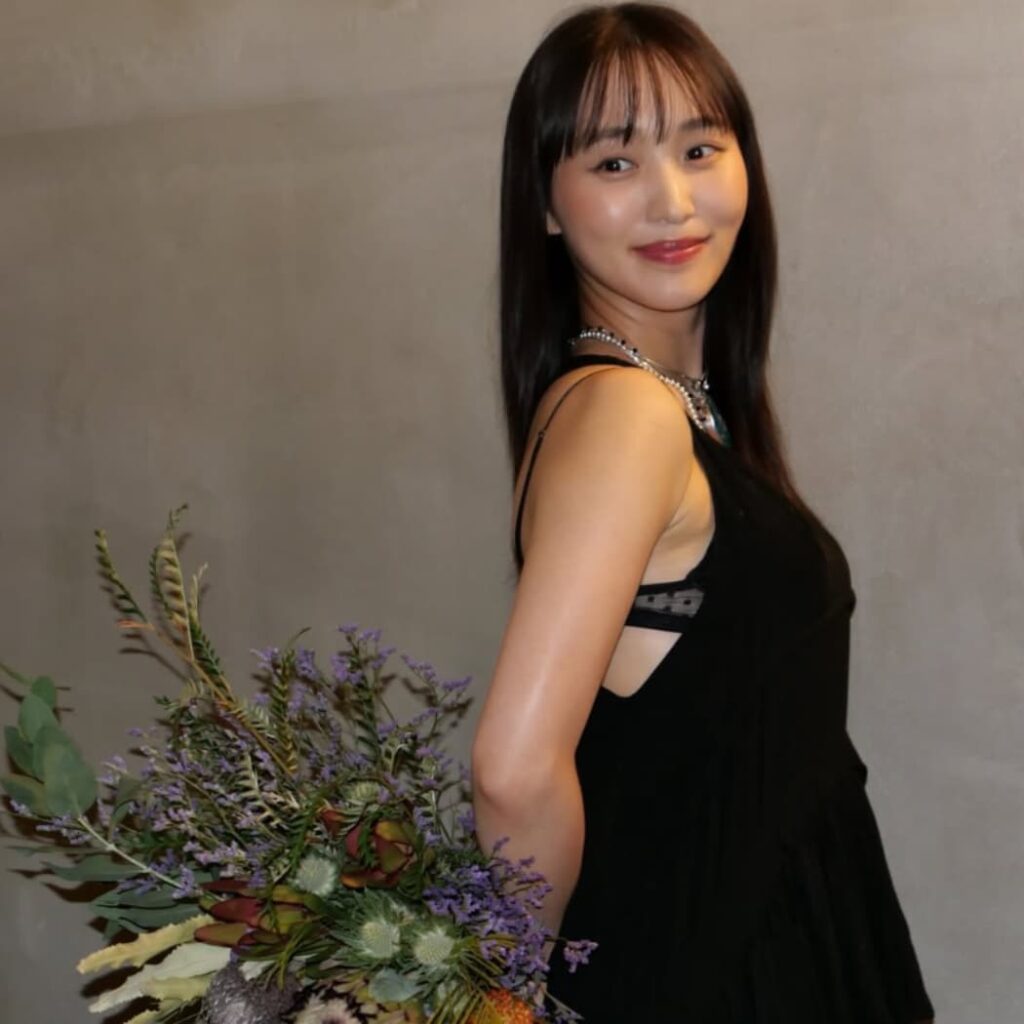 背中の開いた黒いドレスを着用し、プロテアやラベンダーカラーの花々を束ねた大きな花束を抱えながら、肩越しに微笑む菅井友香。