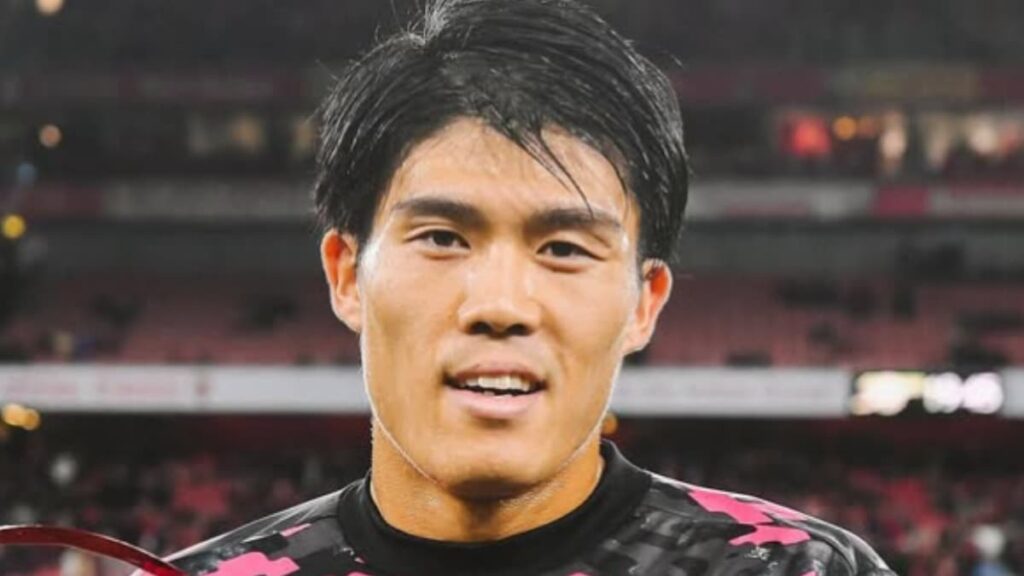 エミレーツ・スタジアムの観客席を背景に、黒いトレーニングウェアを着用して笑顔を見せる冨安健洋のクローズアップ写真。