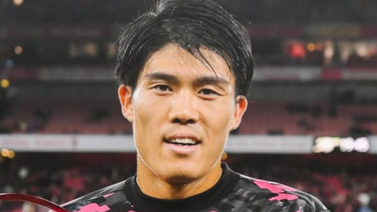 エミレーツ・スタジアムの観客席を背景に、黒いトレーニングウェアを着用して笑顔を見せる冨安健洋のクローズアップ写真。