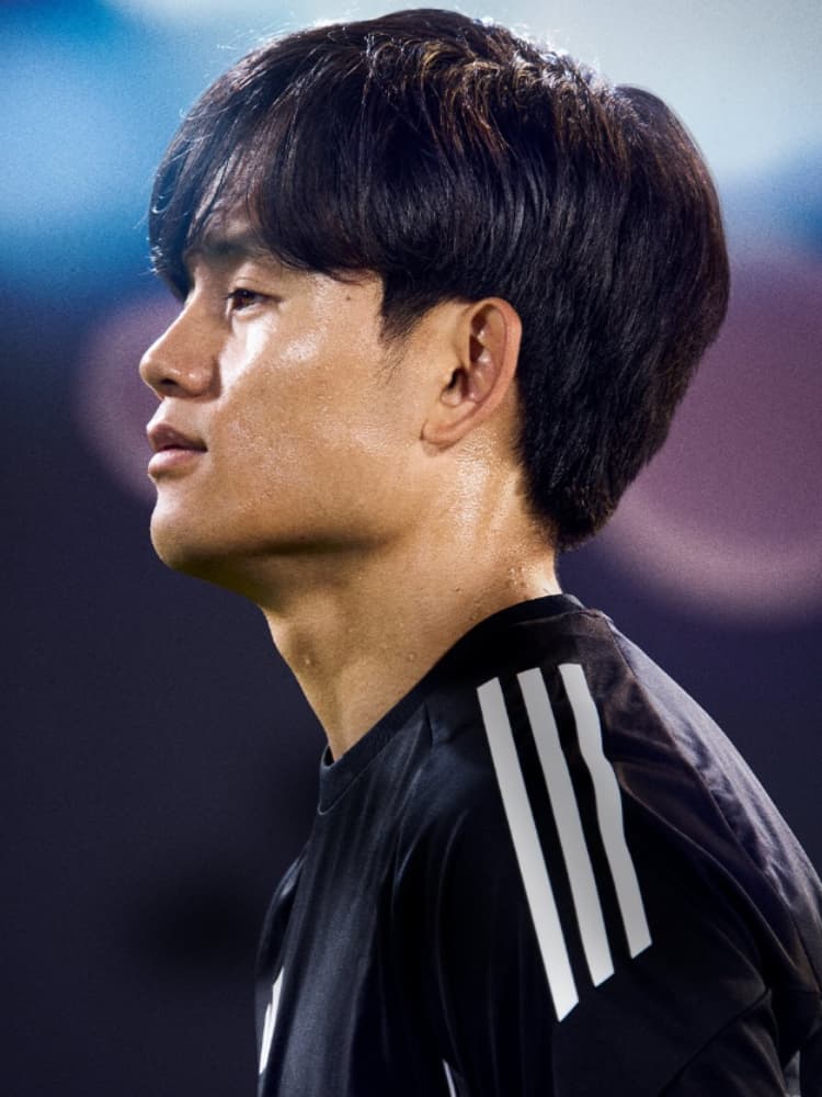 汗をかいた黒いアディダスの練習着を着用し、左向きの横顔を見せる久保建英のクローズアップ。スタジアムの照明が背景にある。