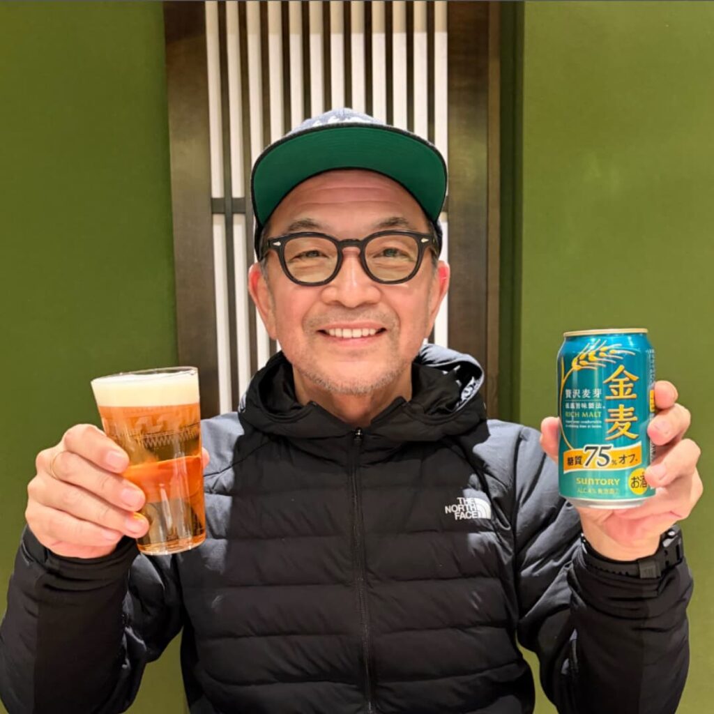 眼鏡と黒い「The North Face」のダウンジャケット、緑のつばの野球帽を着用した中野英雄氏が、緑の壁と格子戸を背景に、笑顔でビールが注がれたグラスと「金麦」の缶を両手に持っている写真。
