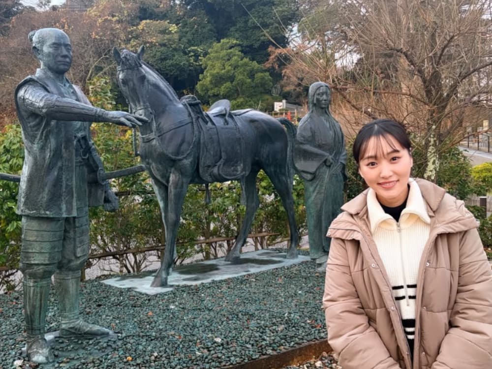 ベージュのダウンジャケットを羽織り、前田利家公とまつ、そして馬のブロンズ像の前で冬の装いをして微笑む菅井友香。