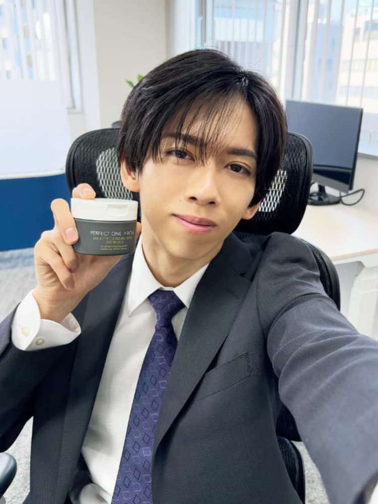 オフィスのデスクで、右手に白いジャー「PERFECT ONE FOCUS SMOOTH CLEANSING BALM」を持ち、カメラに提示する船津稜雅。黒いスーツに青と紫のネクタイを着用。