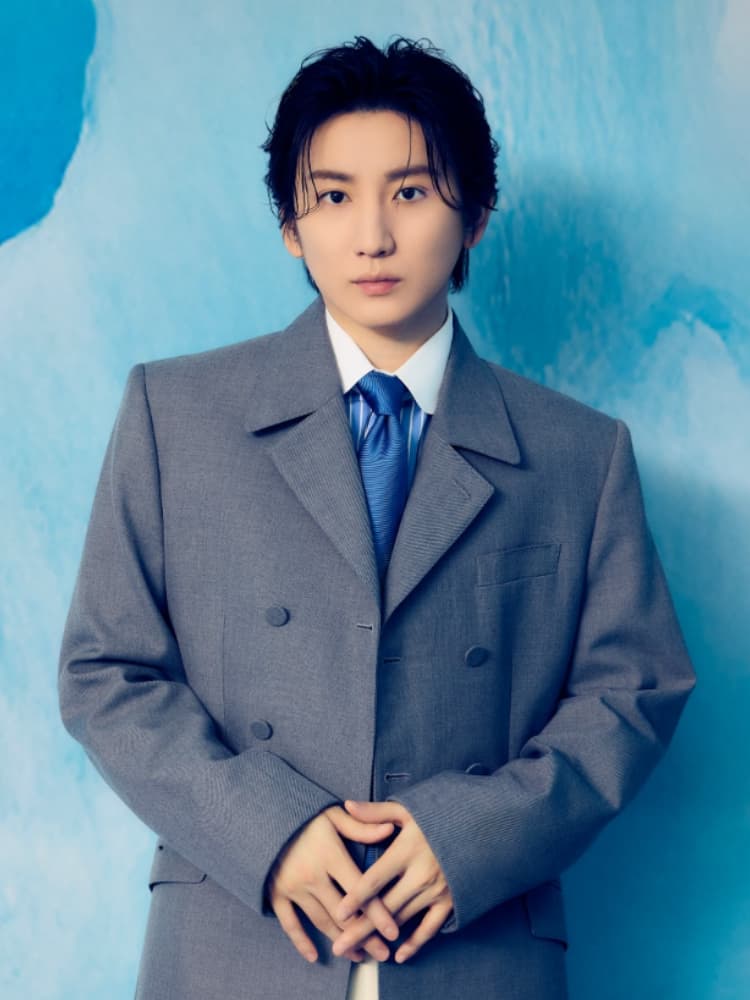 青い水彩風の背景で、グレーのダブルブレストスーツ、青と白のストライプシャツ、青いネクタイを着用した京本大我。両手を前で組んでいる。