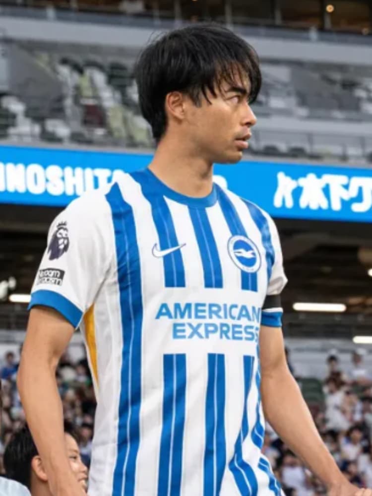 ブライトンのホームユニフォーム（白と青のストライプ）を着用し、スタジアムで前を見据える三笘薫選手。