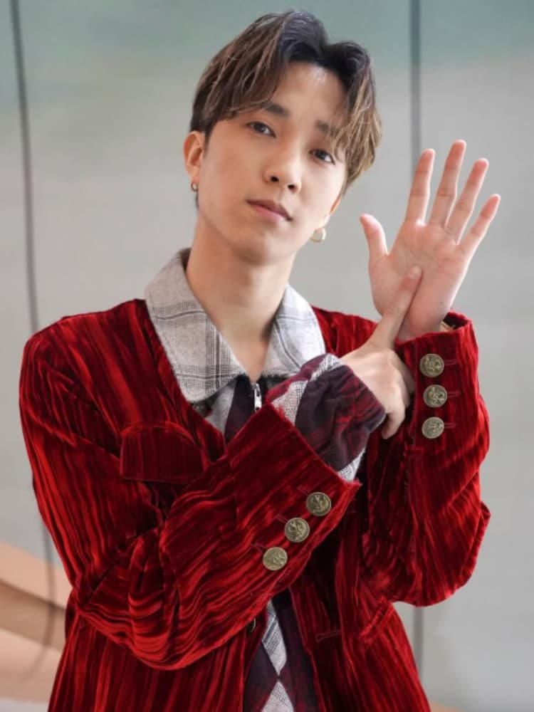 茶髪のミディアムショット。田中樹は赤いベルベットのジャケットとチェックのシャツを着用し、ゴールドのフープイヤリングをつけている。カメラを真剣な表情で見つめながら、右手のひらを広げ、左手の人差し指でその中心を指差すポーズをとっている。このポーズは、SixTONESの象徴的なグループポーズであり、彼らの核心的なコンセプトである「6つの音色（6人のメンバー）の結束」を視覚的に表現している。具体的には、広げた右手の全体像（手のひら）を「6」と見立て、その中心を左手の人差し指で指すことで、6人の多様な『音色』が1つに合わさる（ユニゾンする）という意味を象徴している。背景は明るいグレーの壁。